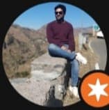 Man in maroon sweater and sunglasses sits on a stone wall overlooking scenic mountains.