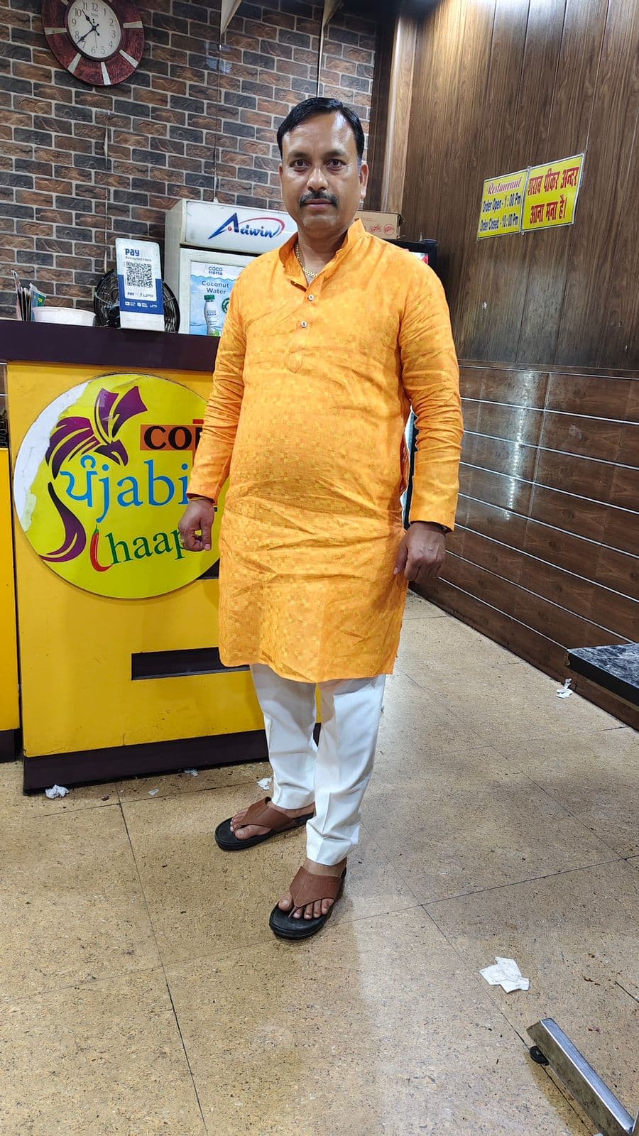 Man in orange kurta and white trousers stands before a yellow Punjabi Chaap food counter.